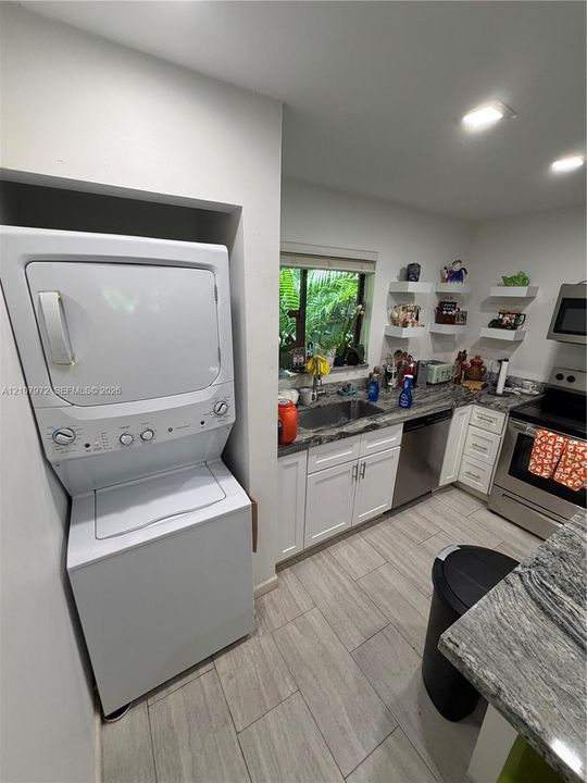 Kitchen featuring white shaker cabinetry, stone-patterned countertops, stainless steel appliances, wood-finish tile flooring, and integrated stacked laundry unit