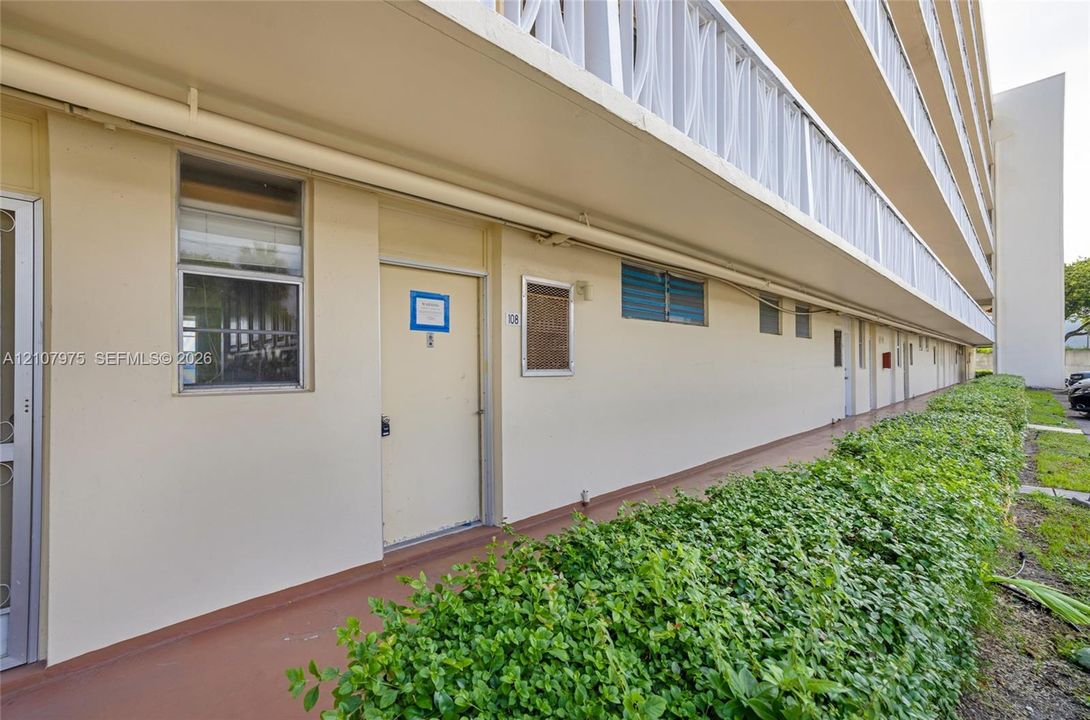 Exterior walkway featuring a covered balcony, recessed window framing, solid-surface flooring, and mature hedge landscaping