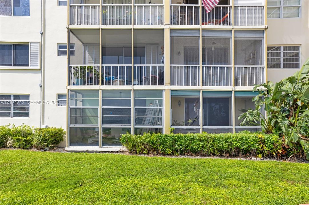 Multi-story building exterior featuring screened-in lanais, aluminum-frame balcony railings, and exterior windows with horizontal sliding panes, situated on a manicured lawn with mature landscaping