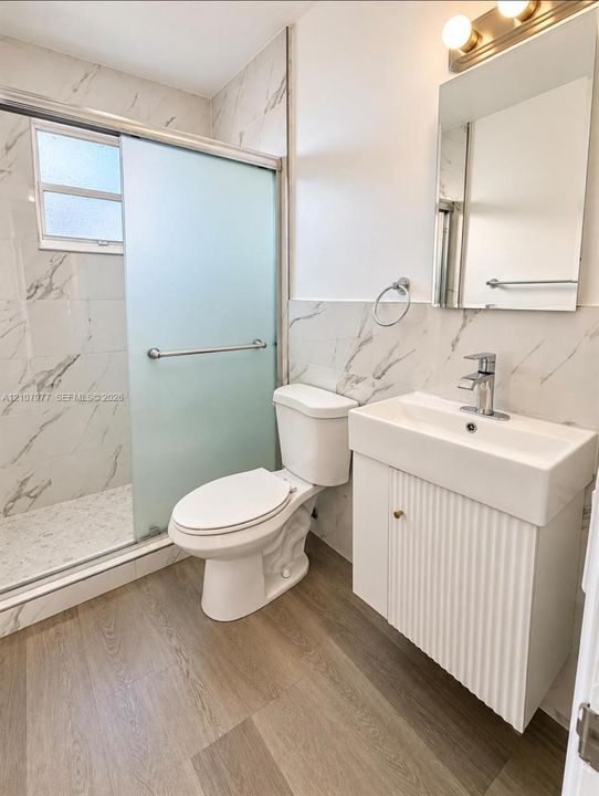 Bathroom featuring stone-finish wall tile, fluted-front vanity with a rectangular sink, walk-in shower with frosted sliding glass doors, and wood-finish flooring