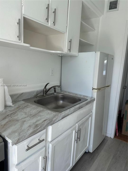 Kitchenette featuring white shaker-style cabinetry with satin nickel hardware, stone-patterned laminate countertops, stainless steel sink with high-arc faucet, and wood-finish flooring