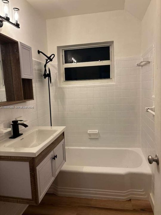 Wall-mounted vanity with wood-finish detailing, white ceramic sink, matte black faucet, full-height white tile tub surround, and integrated wall lighting