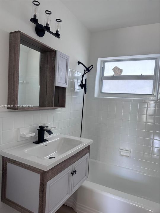 Bathroom featuring a wood-finish vanity with a rectangular sink, matte black faucet, wall-mounted three-light fixture, and white tiled wall surround