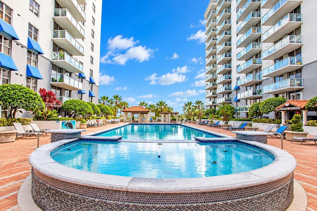 Outdoor swimming pool featuring a tiled spa with a stone-accented exterior, expansive terracotta-toned deck, shaded cabanas, and mature landscaping