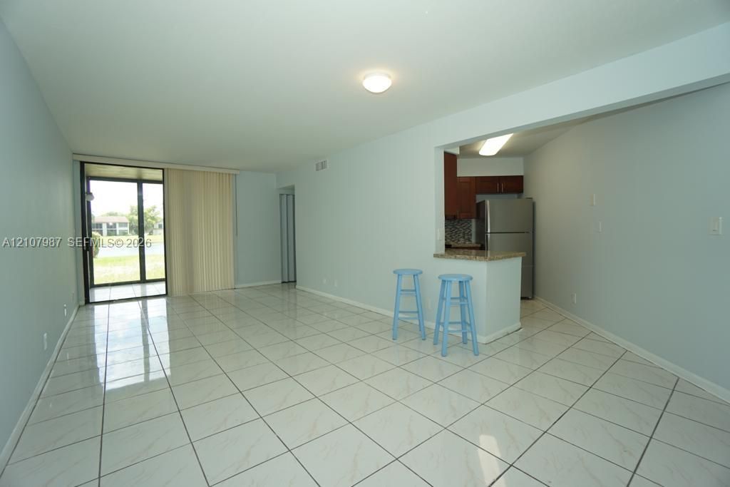 Open-concept interior featuring light-toned tile flooring, pass-through kitchen counter with stone-finish surface, stainless steel refrigerator, and glass sliding doors