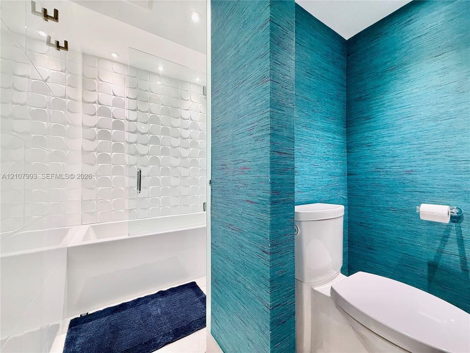 Textured teal wall covering, geometric white tiled shower surround, glass shower partition, and white dual-flush toilet