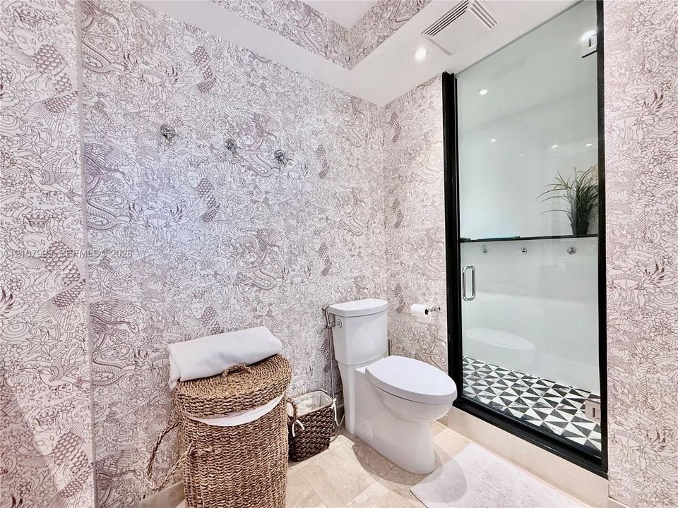 Patterned wall covering, floor-to-ceiling glass shower enclosure with matte black frame, geometric tile shower flooring, and two-piece toilet