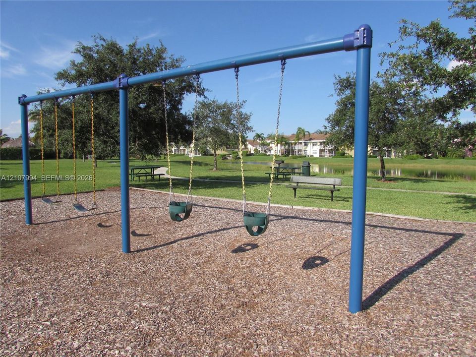 Outdoor recreational area featuring a metal swing set, wood-chip ground cover, and built-in benches situated on a manicured lawn