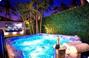 Outdoor spa area featuring a built-in hot tub, perimeter privacy hedging, mature palm trees, and an exterior wall-mounted display screen