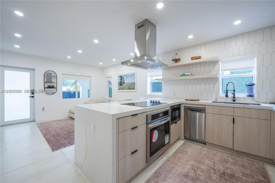 Modern kitchen featuring light-tone wood cabinetry, white stone-finish countertops, stainless steel appliances, a high-reach professional style faucet, and geometric tile backsplash with integrated shelving