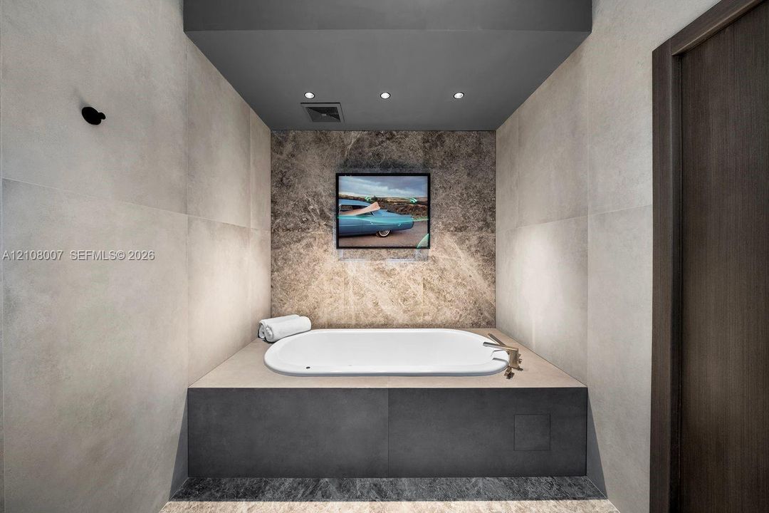 Built-in drop-in bathtub featuring a dark stone-finish base, large-format matte wall tiles, natural stone accent wall, and recessed ceiling lighting