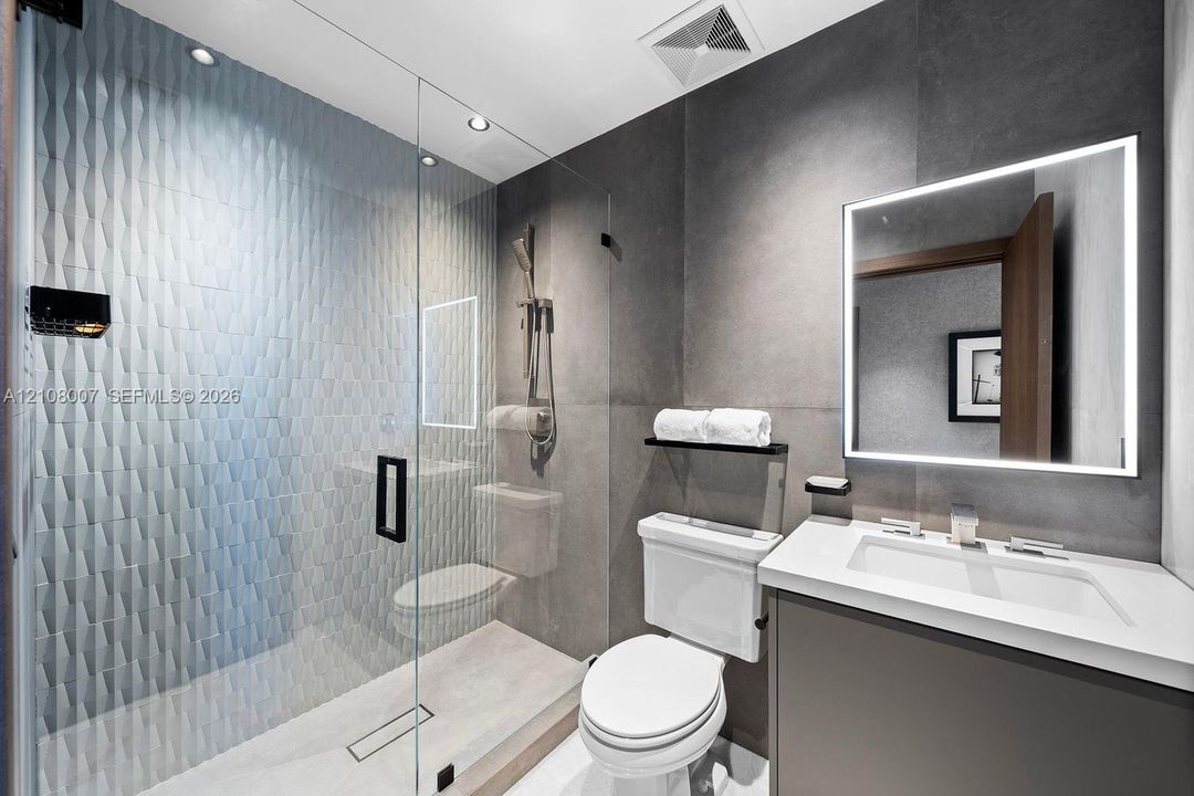 Walk-in glass-enclosed shower featuring three-dimensional tile accent wall, vanity with solid surface countertop, integrated LED-backlit mirror, and matte black fixtures