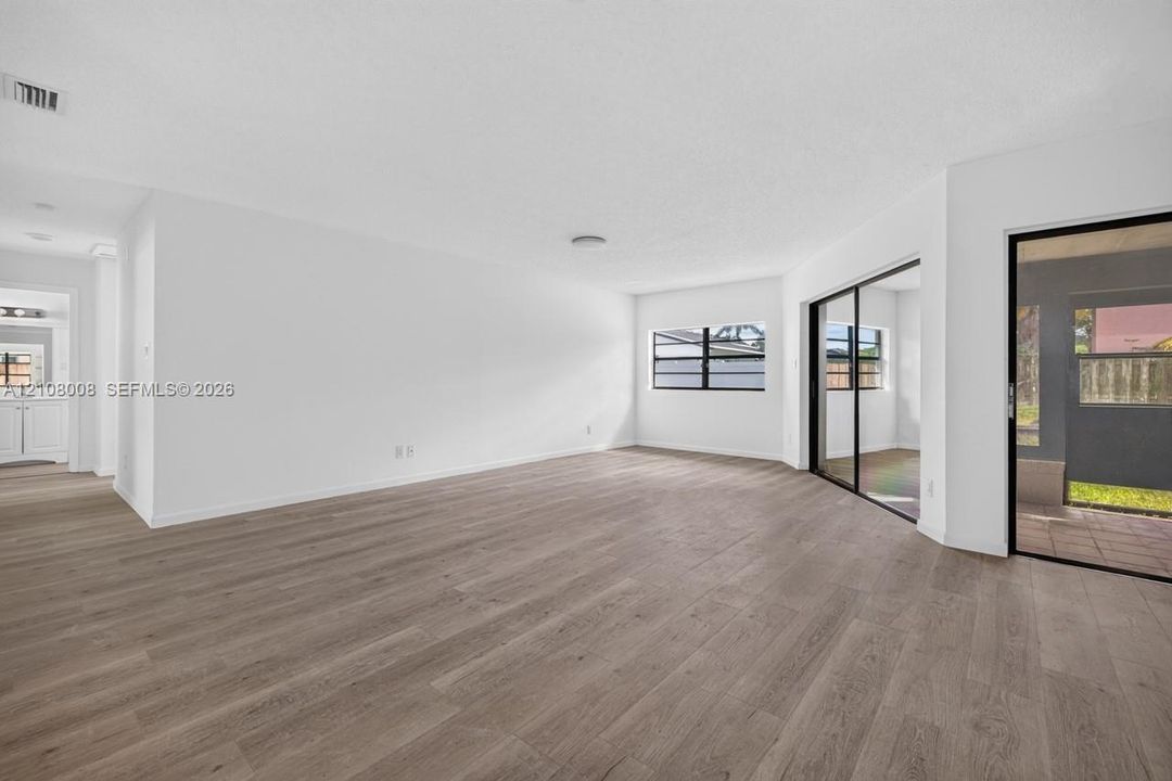 Open-concept area featuring wood-finish flooring, white walls, black-framed windows, and sliding glass doors with access to an exterior screened enclosure