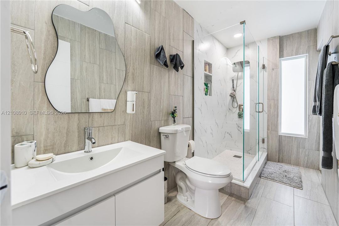 Bathroom featuring vertical wood-finish wall tile, white vanity with an integrated sink, frameless glass-enclosed shower with a recessed shelf, and an irregular-shaped wall mirror