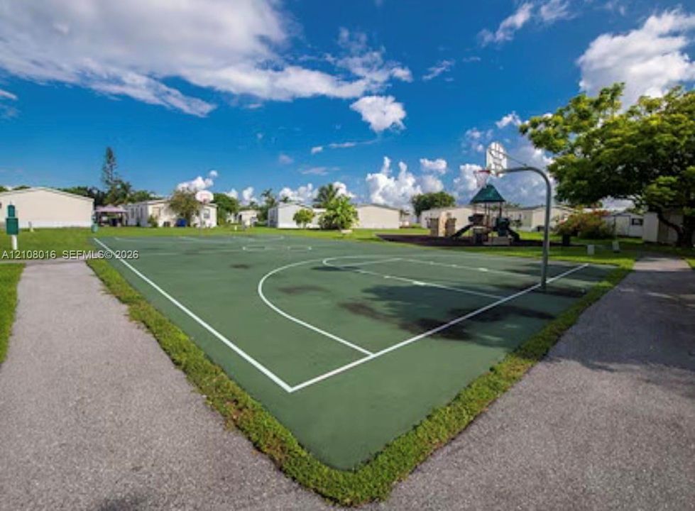 Outdoor basketball court with a paved surface, metal hoop assembly, and supplemental playground structure situated within a landscaped common area