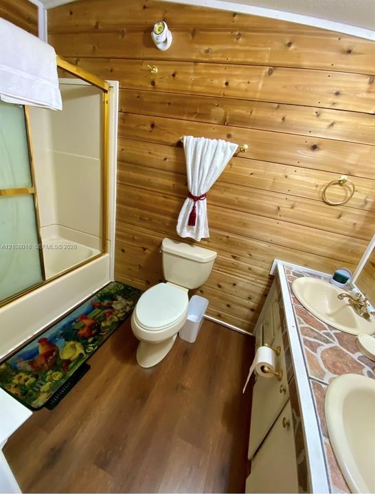 Bathroom featuring wood-paneled walls, wood-finish flooring, a dual-sink vanity with a stone-tile countertop, and a shower-tub combination with a sliding enclosure