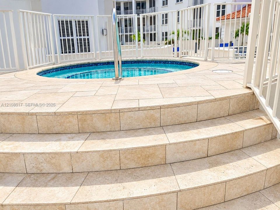 Elevated spa featuring a blue tiled interior, metal handrail, surround of stone-finish paver decking, and white perimeter fencing