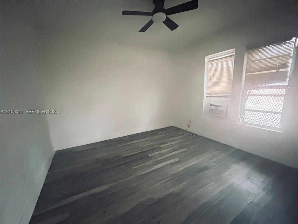 Interior room featuring wood-finish flooring, a multi-blade ceiling fan with central lighting, dual windows with blinds, and an integrated wall-mounted air conditioning unit