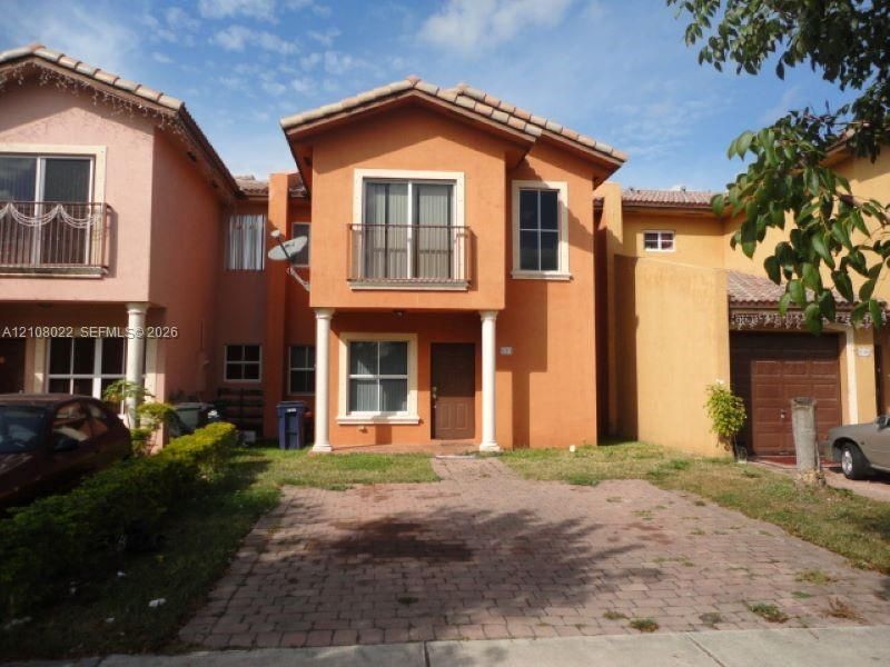 Stucco exterior featuring a paver driveway, arched entry with dual columns, a second-story balcony with metal railing, and a tile roof