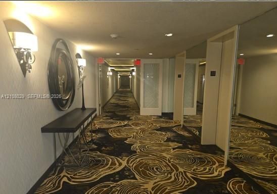 Hallway featuring patterned wall-to-wall carpeting, decorative wall-mounted sconces, textured wall coverings, and mirrored paneling