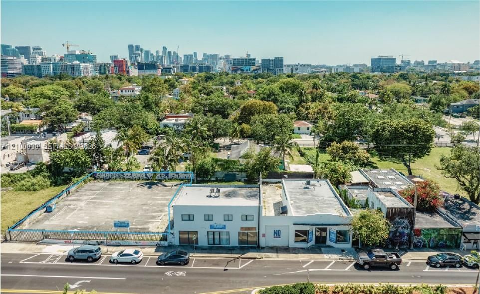 Aerial perspective featuring street-front commercial buildings, paved surface lot, and distant urban skyline backdrop