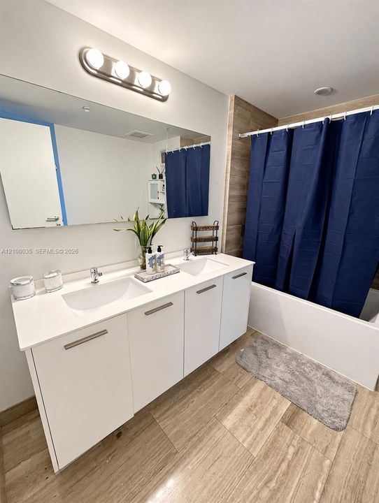 Dual-sink vanity with a solid-surface countertop, full-width vanity mirror, four-light wall fixture, and tub-shower combination with floor-to-ceiling tile surround