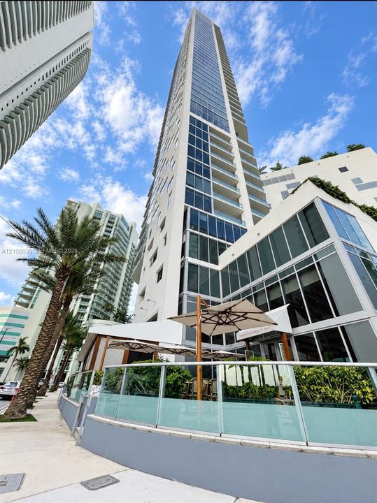 High-rise residential tower featuring a glass and concrete facade, wrap-around balcony levels, and an expansive ground-level terrace with integrated glass railings