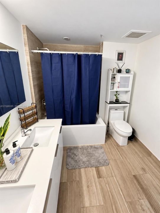 Modern bathroom featuring wood-finish tile flooring, a dual-sink vanity with a solid surface countertop, and a shower-tub combination with wall-to-wall tiling