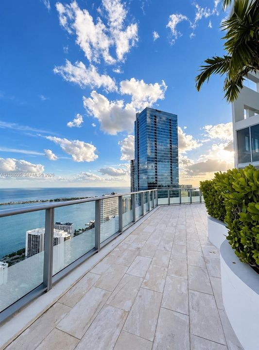 Expansive terrace featuring large-format stone tiling, a glass panel railing system, and built-in curved planters with mature shrubbery