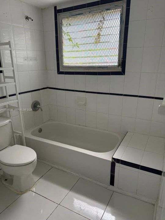 Bathroom featuring white tile flooring and wall tiling with dark accent trim, built-in tiled tub surround, integrated soaking tub, and a window with a security screen