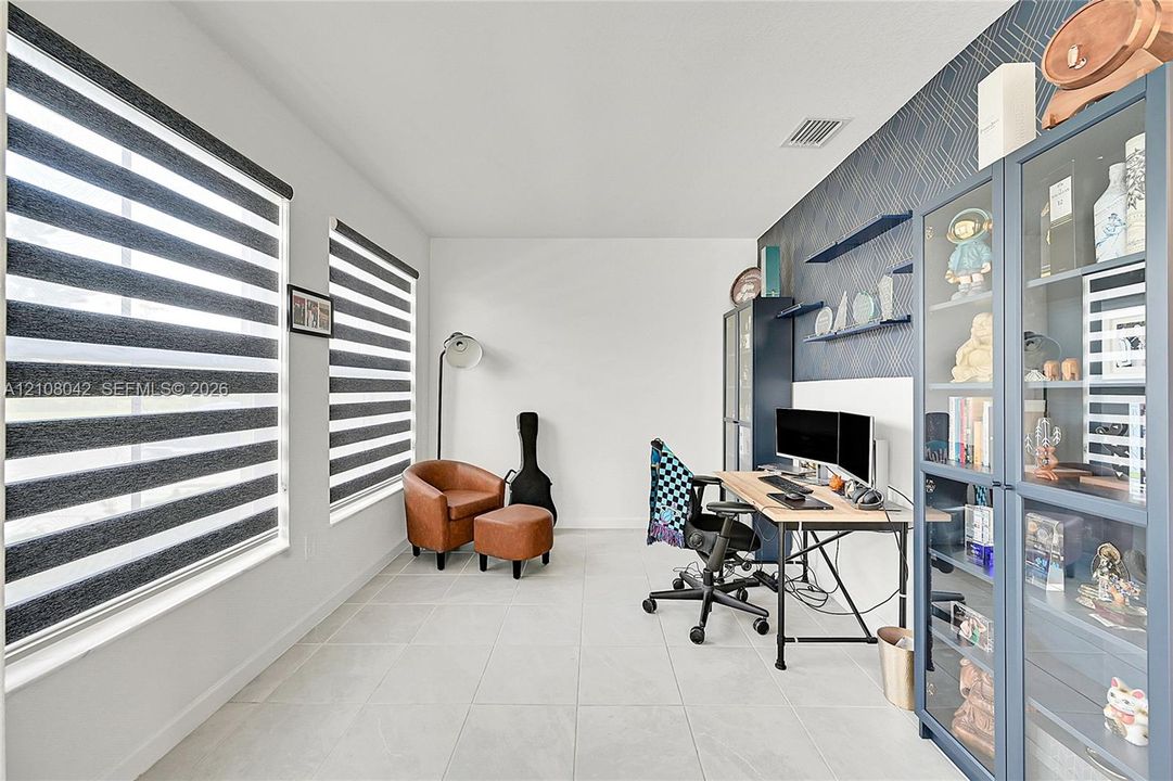 Room featuring neutral tile flooring, dual-layered roller shades, and a geometric-patterned accent wall with integrated shelving