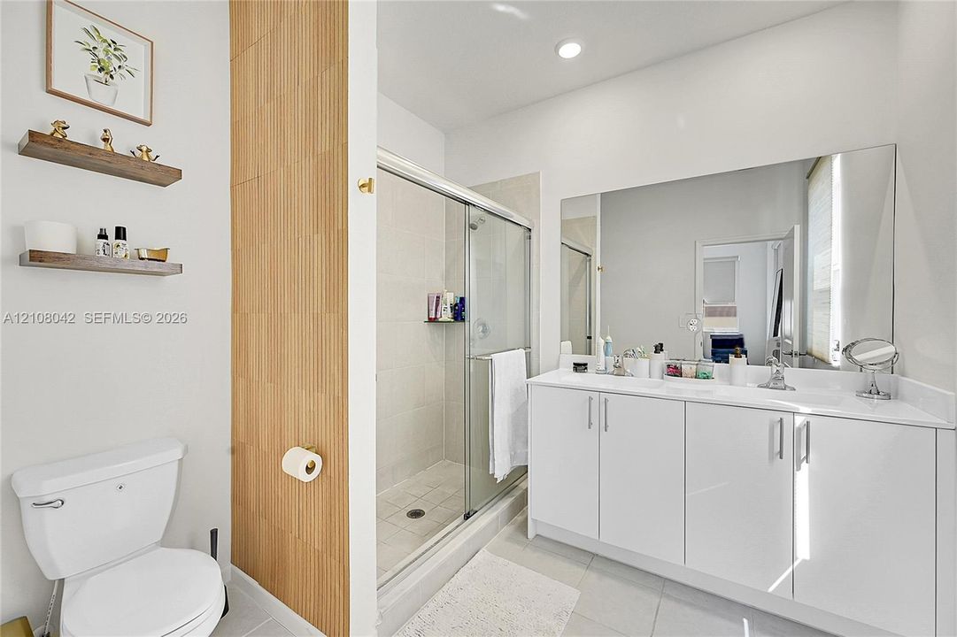 Bathroom featuring a white dual-sink vanity with a solid-surface countertop, floor-to-ceiling wood-panel accent wall, walk-in shower with sliding glass enclosure, and two wall-mounted floating shelves