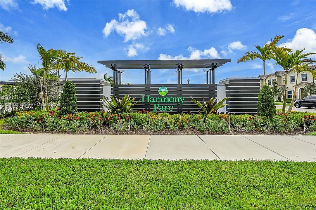 Community entrance monument featuring horizontal slat architectural detailing, integrated signage, concrete walkway, and professionally landscaped tropical foliage