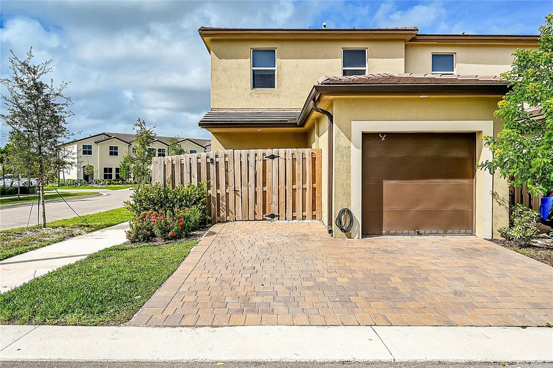 Two-story stucco exterior featuring a paved stone driveway, an attached garage, and a wood privacy fence