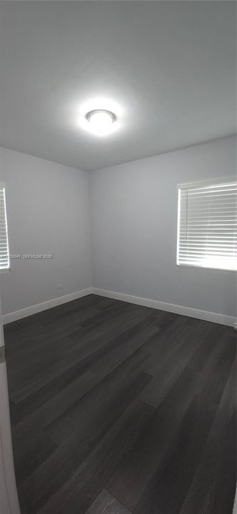 Neutral-toned interior featuring wood-finish flooring, white walls, two windows with horizontal blinds, and a flush-mount ceiling light fixture