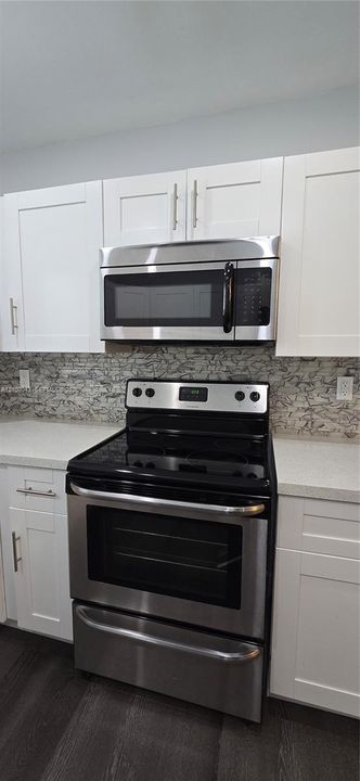 Shaker-style white cabinetry, stainless steel microwave, stainless steel electric range, stone-mosaic backsplash, and solid-surface countertops