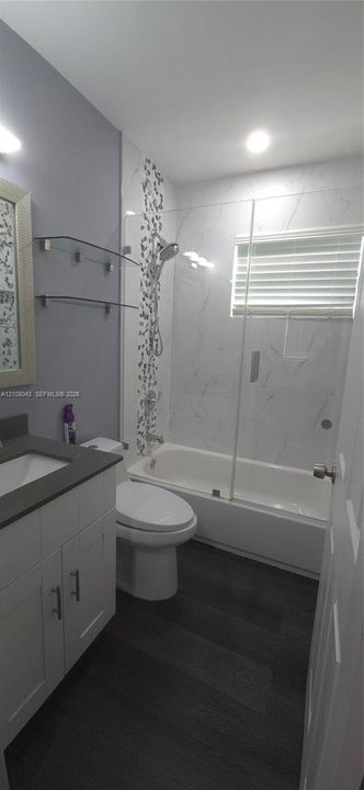 Bathroom featuring a white shaker-style vanity with a dark solid-surface countertop, wood-finish flooring, a combination tub and shower with a glass panel enclosure, and tile wall accents