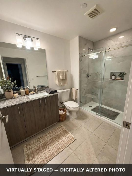 Bathroom featuring wood-finish cabinetry, stone-patterned countertop with an integrated rectangular sink, large format tile flooring, and a walk-in shower with glass sliding doors and a built-in recessed shelf