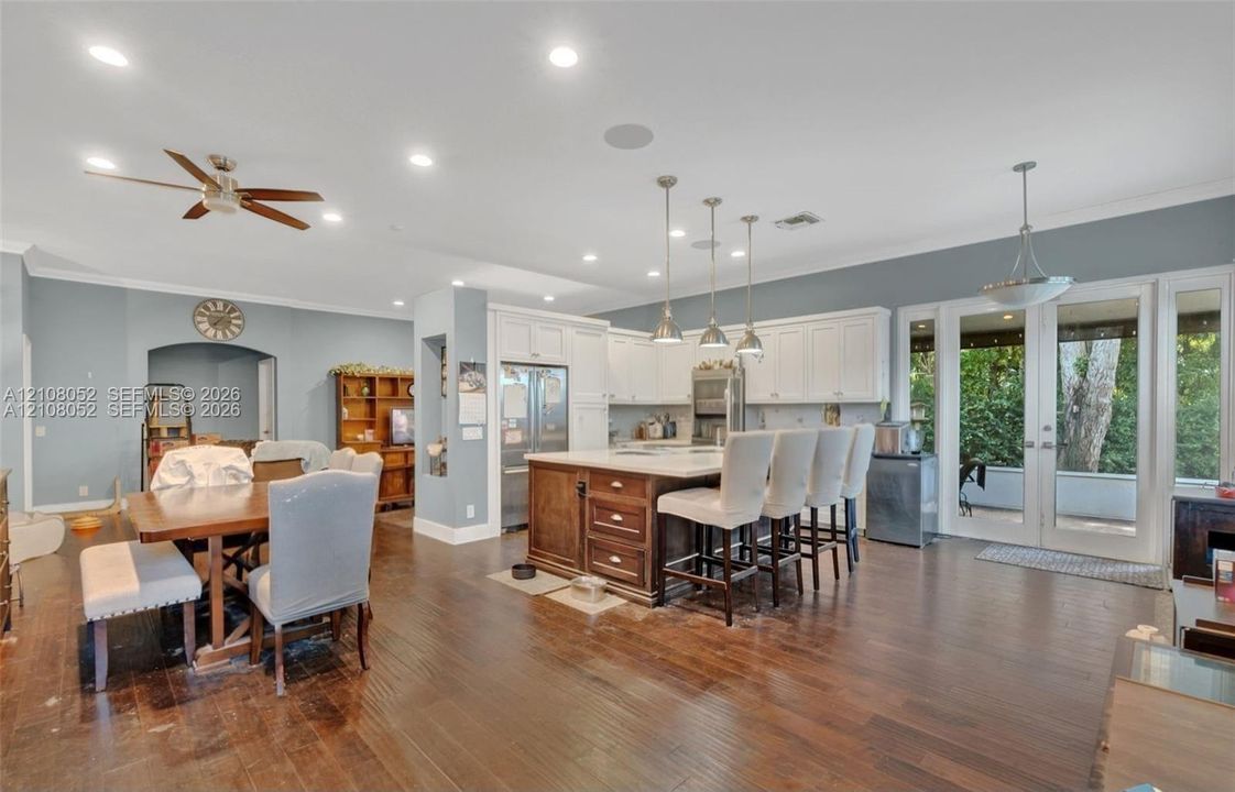 Open-concept kitchen featuring dark wood-finish flooring, a central island with wood cabinetry and stone countertops, white cabinetry, three pendant lights, and French doors leading to a covered patio