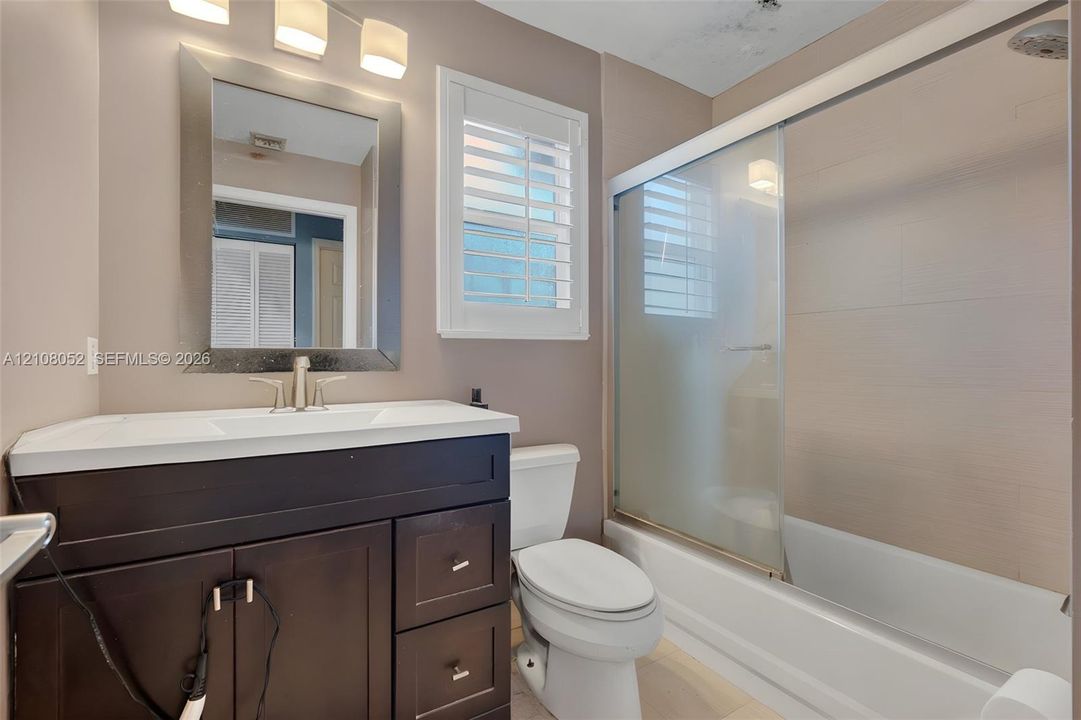 Dark wood vanity with integrated countertop, framed wall mirror, three-light vanity fixture, plantation shutter window covering, and shower-tub combination with sliding glass doors