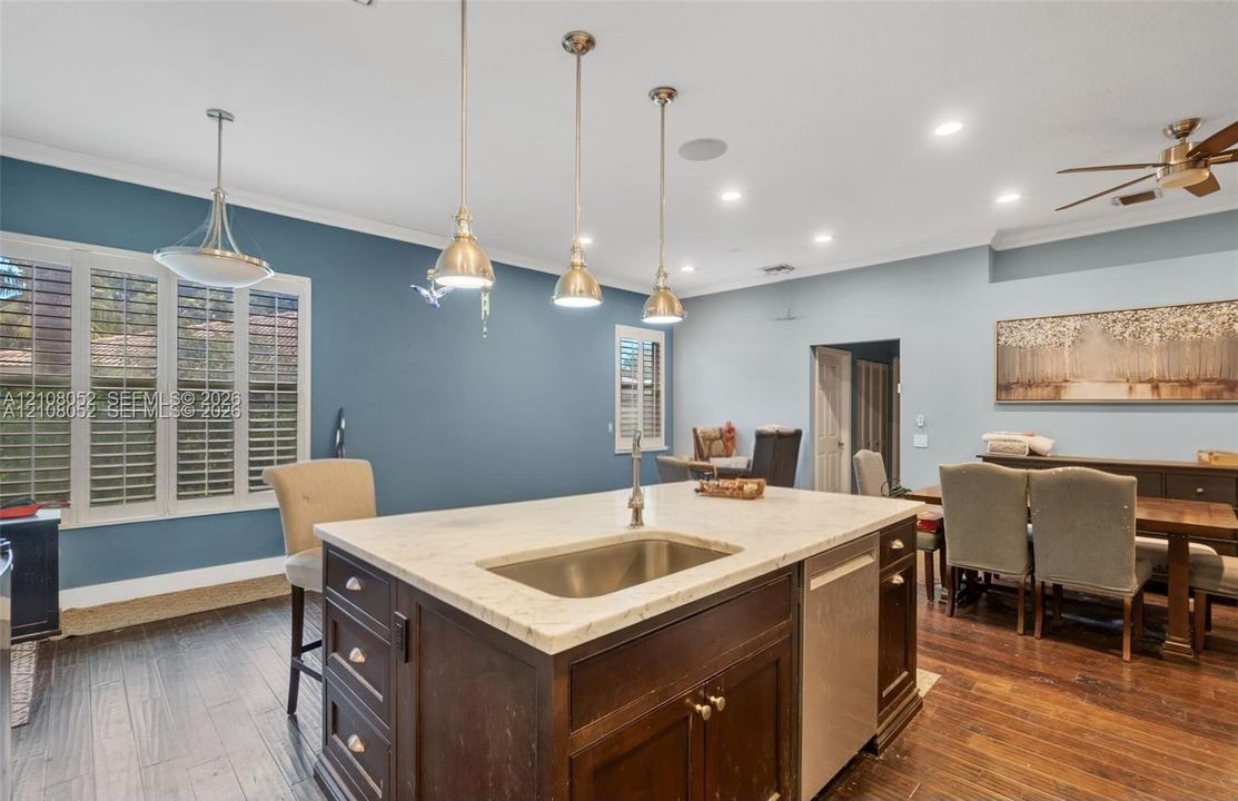 Kitchen island featuring stone countertop, undermount stainless steel sink, and dark wood-finish cabinetry, complemented by wood-finish flooring, recessed lighting, and three pendant lights