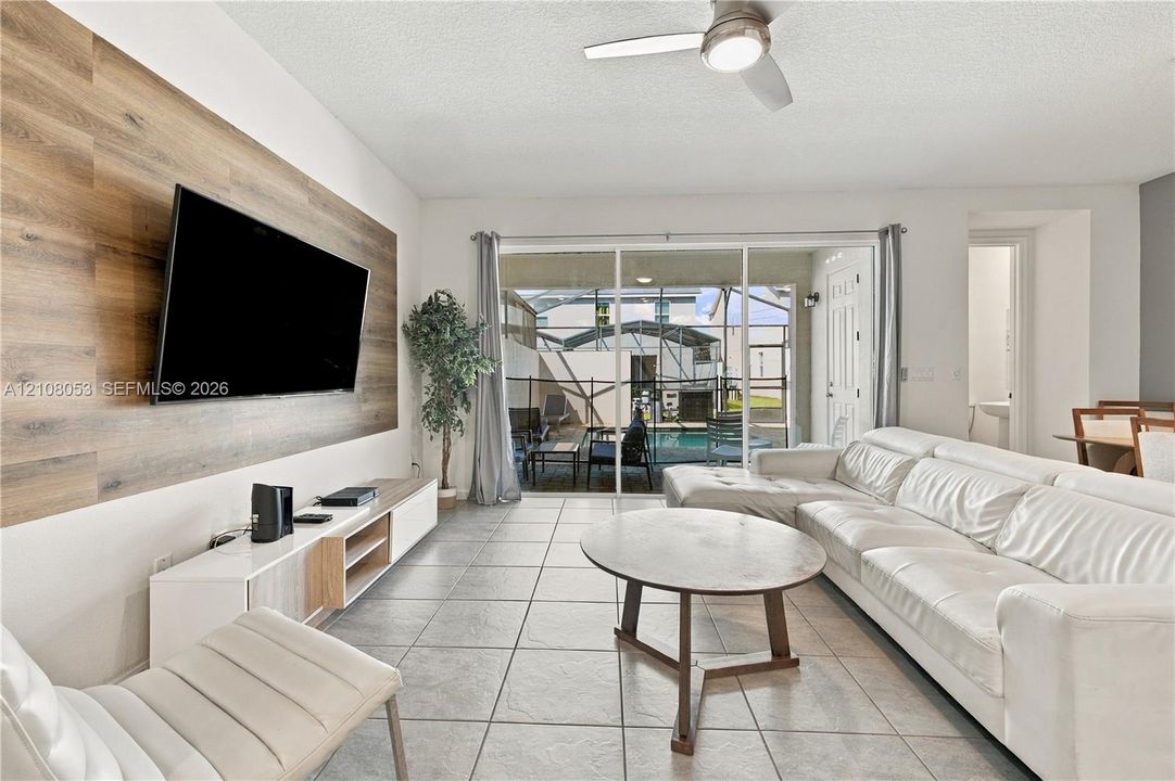 Living area featuring wood-finish accent wall paneling, large-format tile flooring, ceiling fan with integrated lighting, and sliding glass doors providing outdoor access