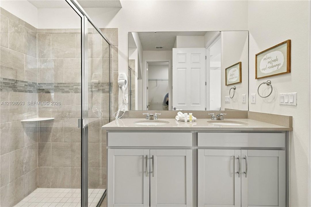 Dual-sink vanity with a solid-surface countertop, full-width mirror, glass-enclosed walk-in shower with tile surround, and inset decorative tile detail