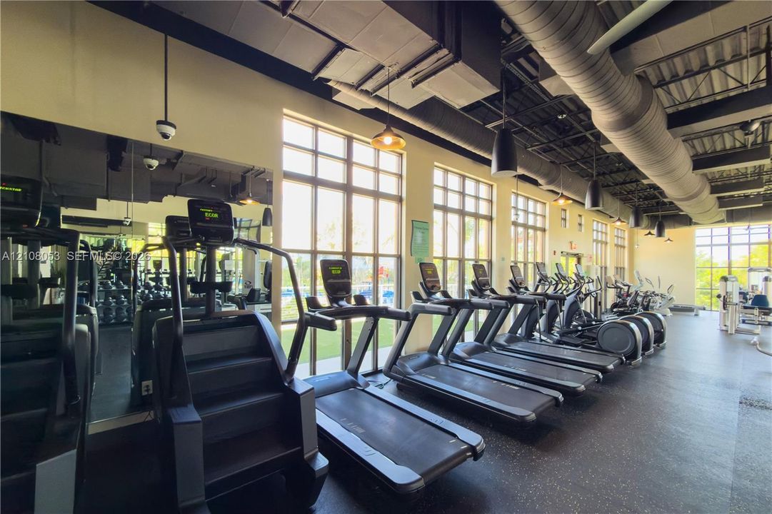 Fitness center featuring floor-to-ceiling windows, high-contrast industrial-style exposed ductwork, wall-to-wall mirrors, and resilient gym flooring