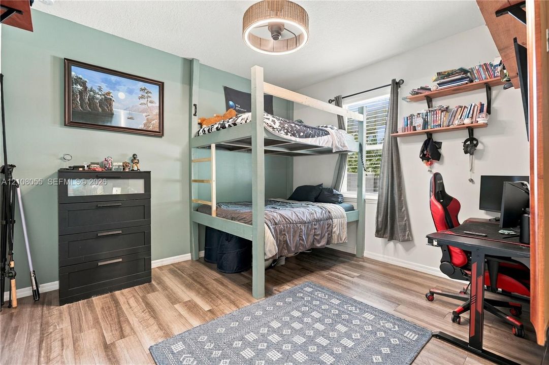 Bedroom featuring wood-finish flooring, a drum-style ceiling light fixture, wall-mounted shelving, and a single window with plantation-style shutters
