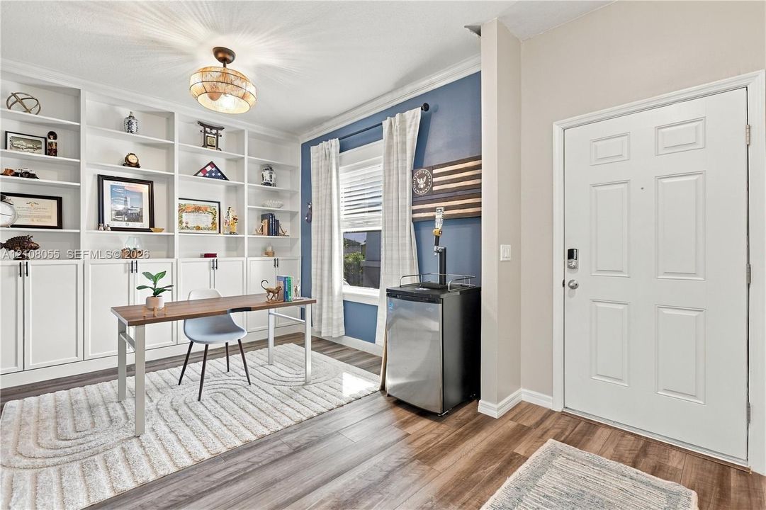 Built-in floor-to-ceiling white shelving with lower cabinetry, wood-finish flooring, multi-pane window with trim, decorative ceiling-mounted fixture, and a six-panel entry door