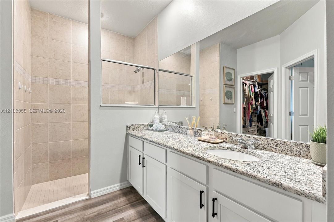 Full-width vanity with stone-finish countertop, white cabinetry with matte black hardware, integrated undermount sink, and tiled walk-in shower with glass enclosure