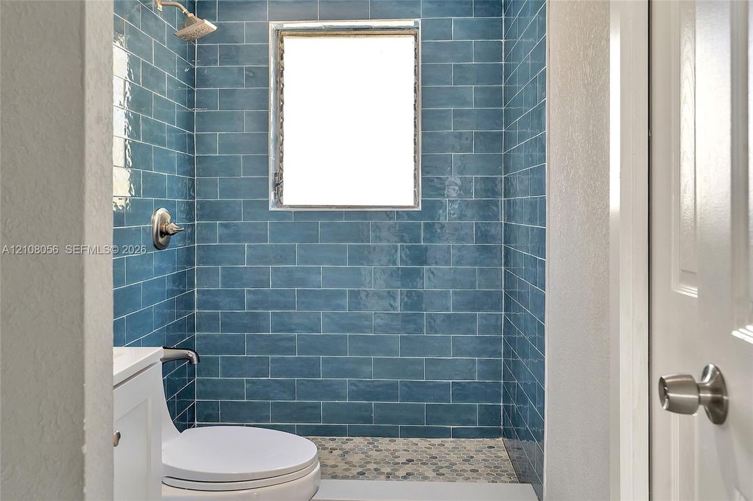 Walk-in shower featuring glossy blue subway tile, a natural stone pebble floor, a built-in shower head, and a privacy window