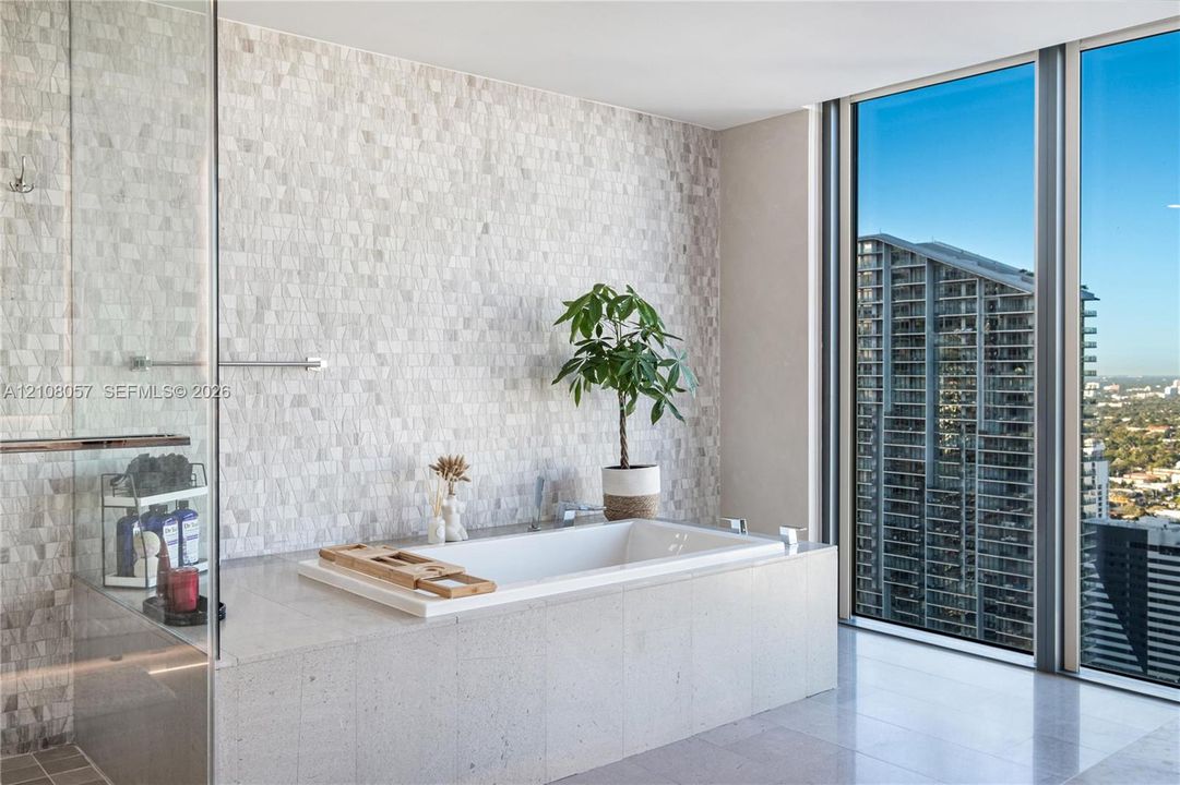 Drop-in soaking tub with a stone-finish surround, floor-to-ceiling windows, mosaic tile wall accent, and a glass-enclosed shower