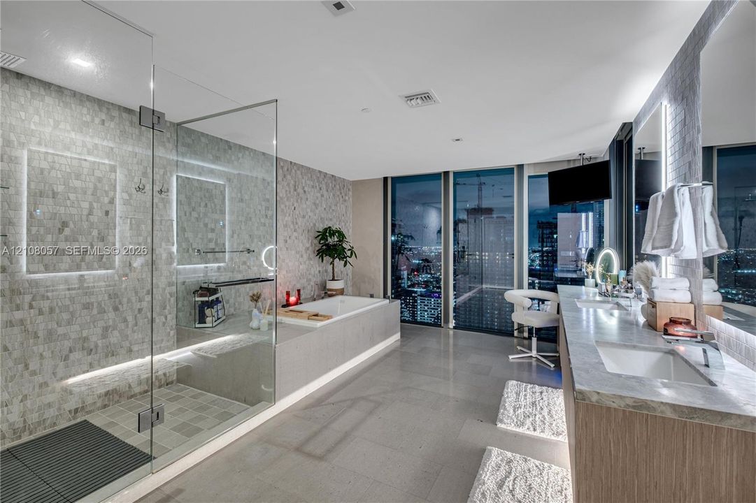 Bathroom featuring floor-to-ceiling windows, a glass-enclosed walk-in shower with stone tile wall, a soaking tub with stone surround, and a dual-sink vanity with stone countertop